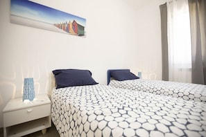 A double bed with a patterned bedspread, a nightstand with a lamp, and a painting of colorful buildings on the wall.