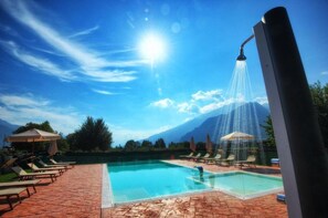 A pool area with lounge chairs and umbrellas, a water feature, and mountains in the background. - Farmhouse at Poncia - Studio Apartment 1 (Gravedona)