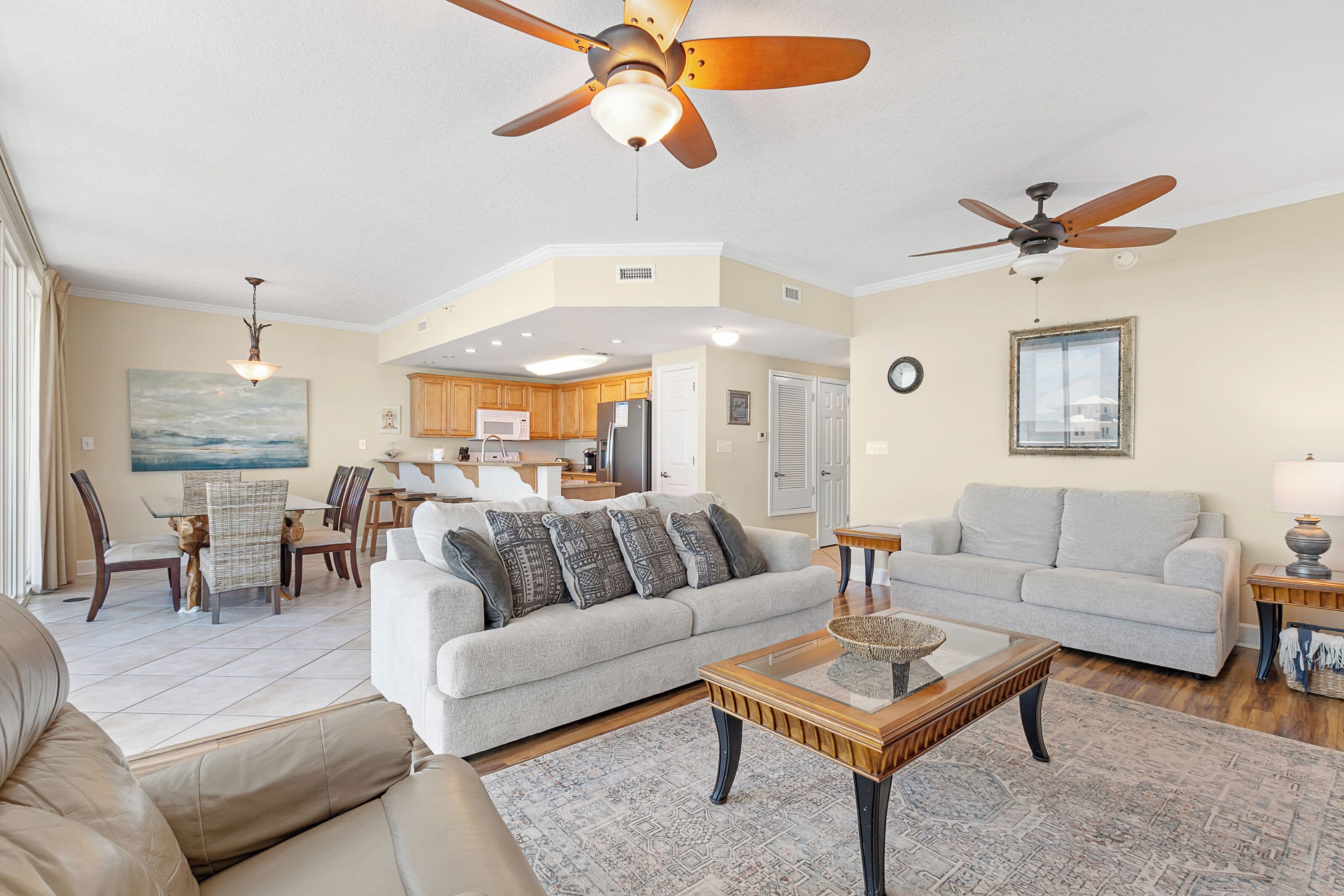 A spacious living room with a large sofa, a coffee table, and a dining area with chairs and a table. There are ceiling fans and a kitchen in the background.