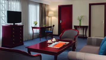 A hotel room with a TV, a dining table with chairs, a sofa, a desk with a chair, and a door.