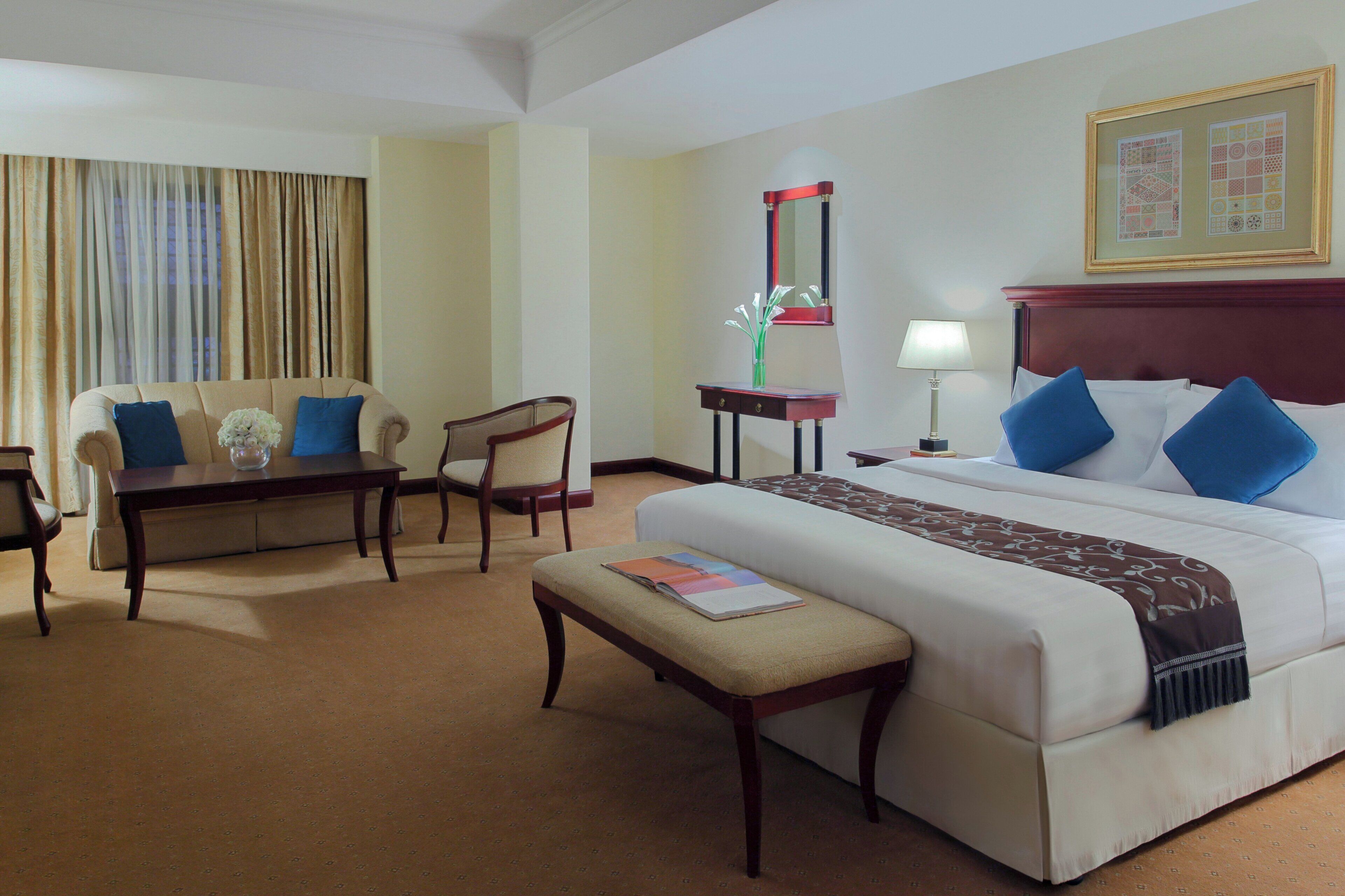 A hotel room with a bed, a desk, a chair, a bedside table, a lamp, and a window with curtains.