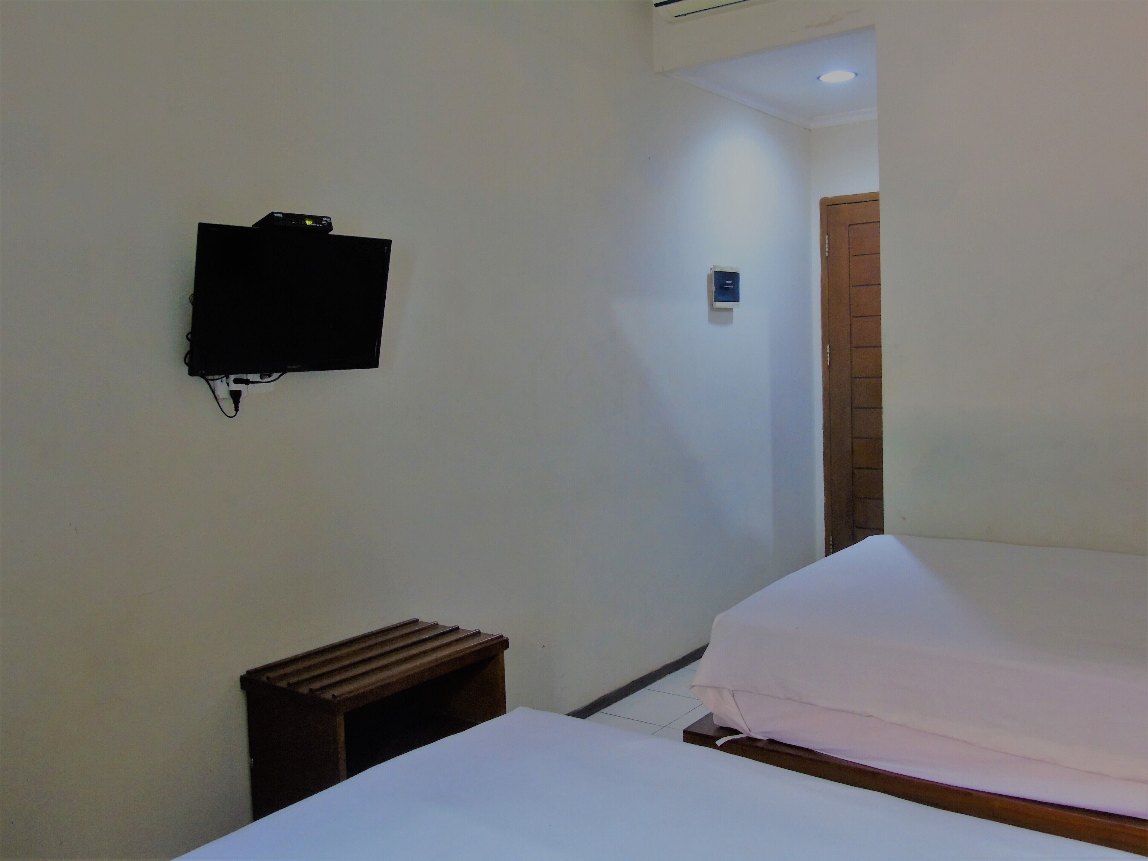 A hotel room with a bed, a small wooden nightstand, a television mounted on the wall, and a door leading to another room.