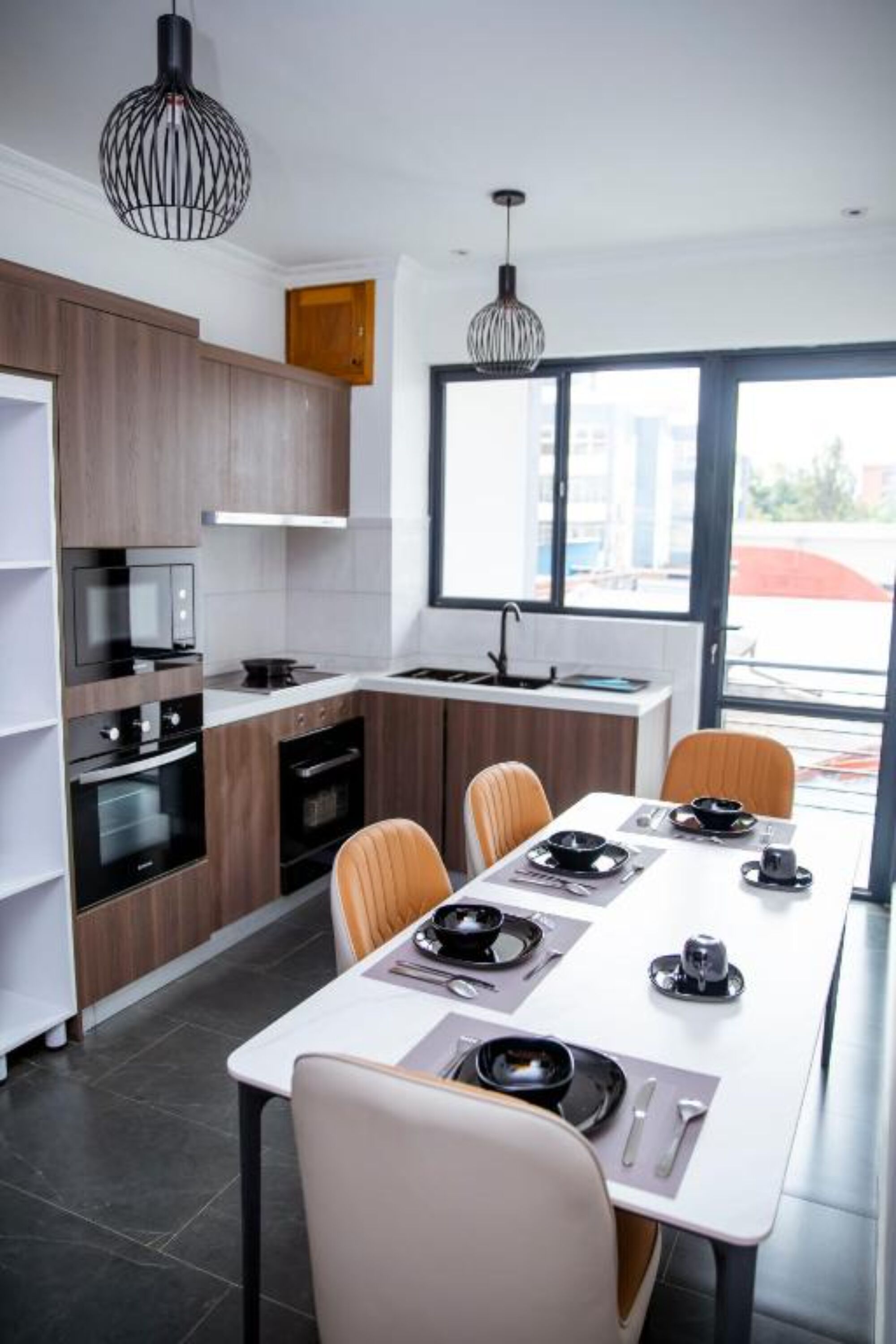 Et moderne køkken med indbyggede apparater, et spiseområde med et hvidt bord og stole, samt store vinduer der giver naturligt lys.