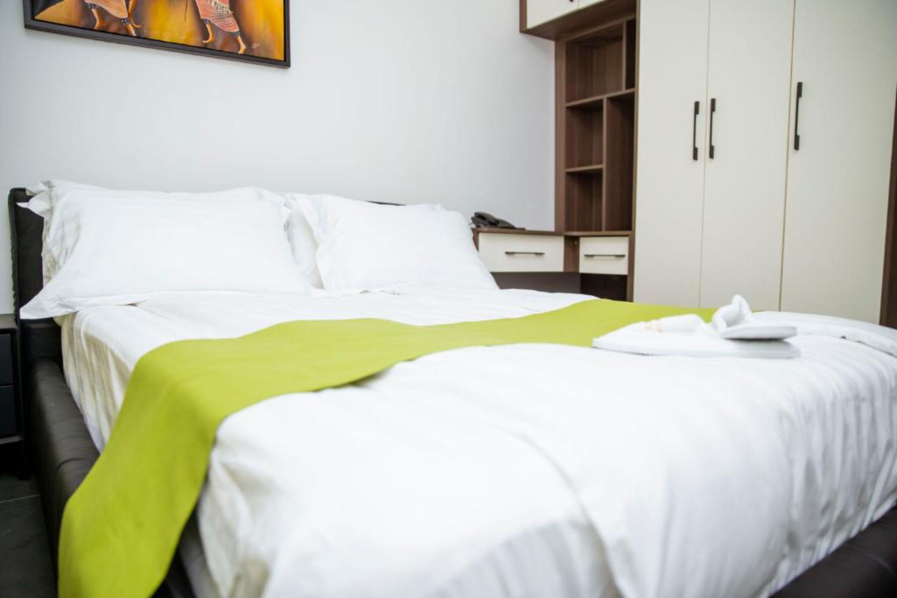 A neatly made bed with white linens and a green throw, a bedside table with a phone, and a wardrobe with multiple doors.