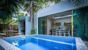 A modern outdoor swimming pool with a patterned tile design, a patio area with a table and chairs, and a kitchen visible through a glass door.