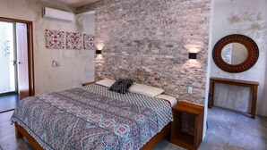 A bedroom with a brick wall, a bed with patterned bedding, two wall-mounted lamps, a round mirror, and a small wooden table.