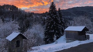 Un chalet rustique aux murs de pierre et à la terrasse en bois, entouré d’arbres couverts de neige et dominé par un paysage montagneux.