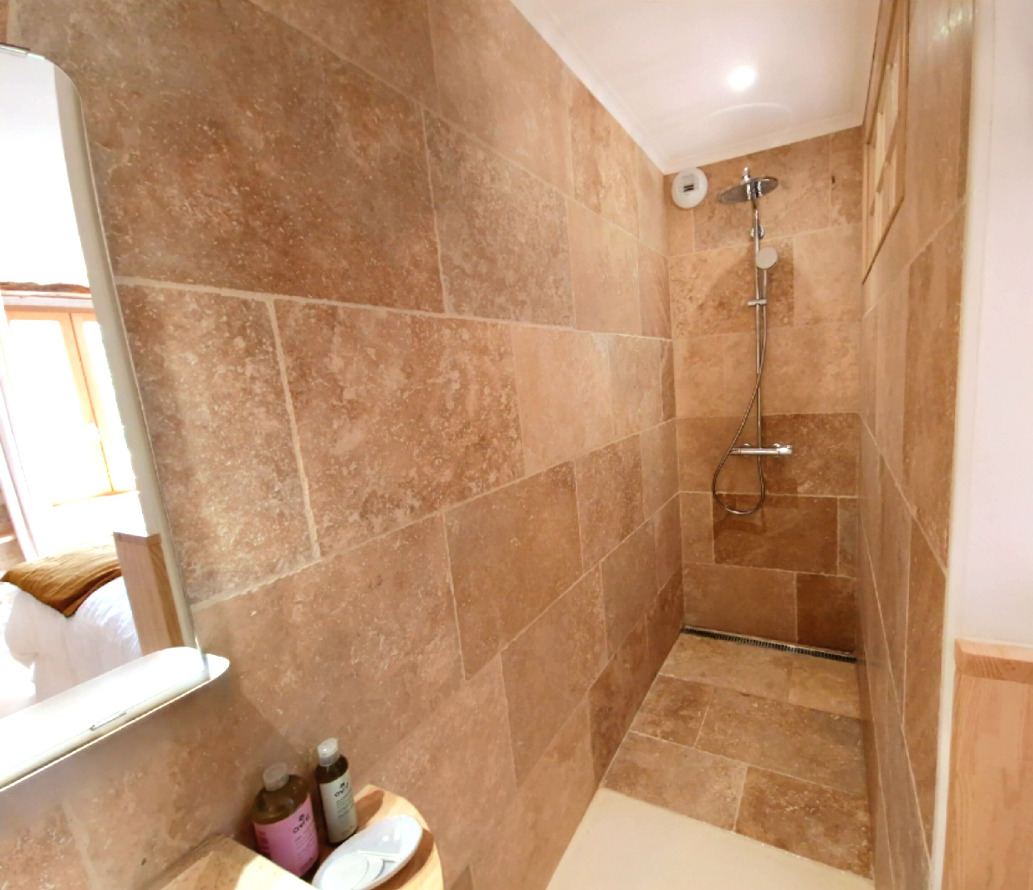 Een badkamer met beige tegels, een douche, een spiegel en een wastafel met toiletartikelen.