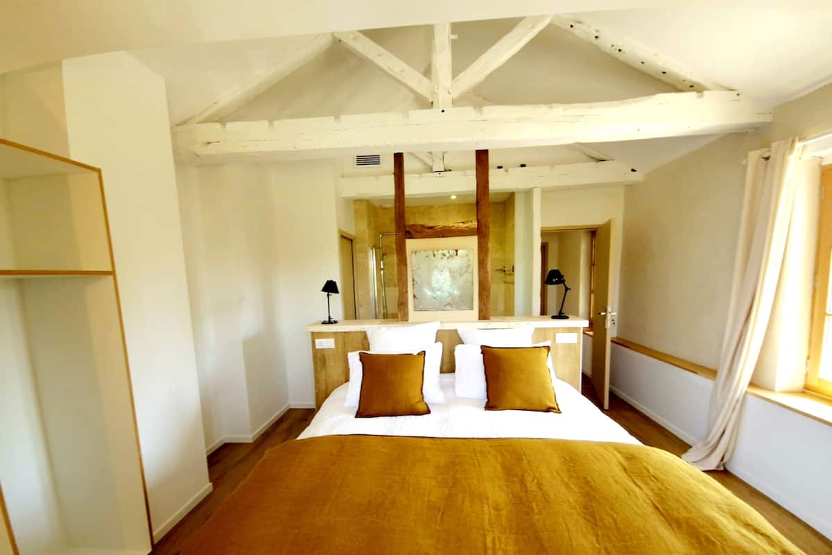 A bedroom with a large bed, a wooden ceiling, a bathroom visible through a glass door, and a window with curtains.