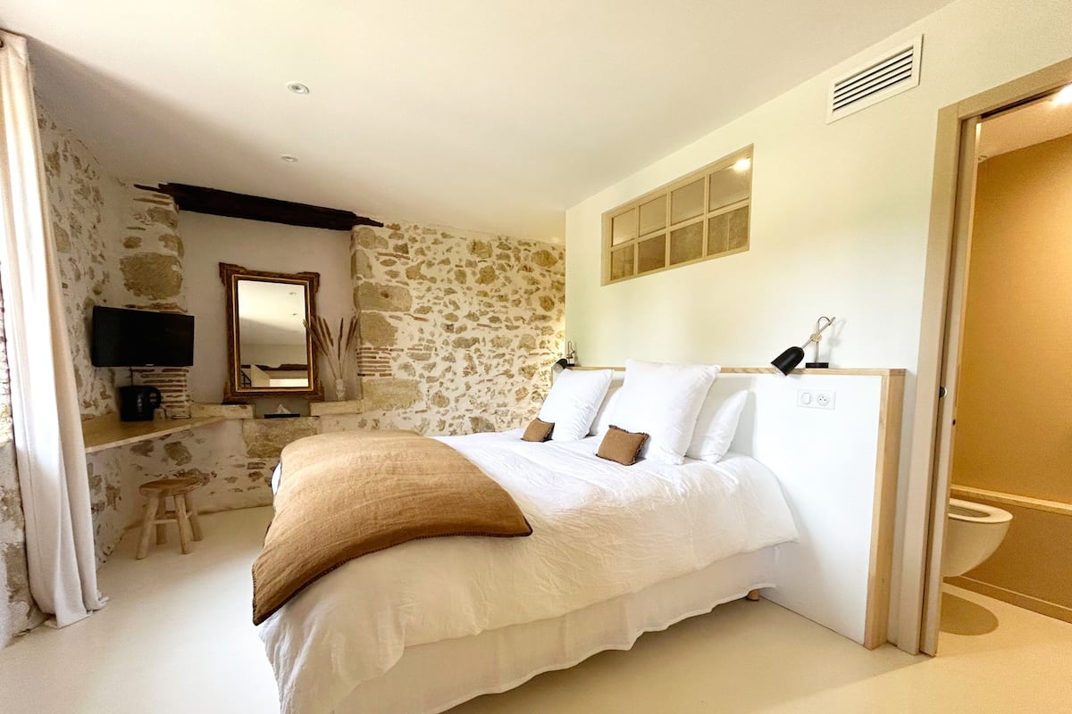 A bedroom with a stone wall, a bed with white bedding, a wooden stool, a television, and a mirror.