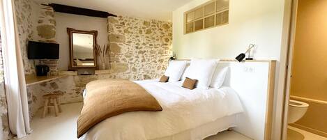 A bedroom with a stone wall, a bed with white bedding, a wooden stool, a television, and a mirror.
