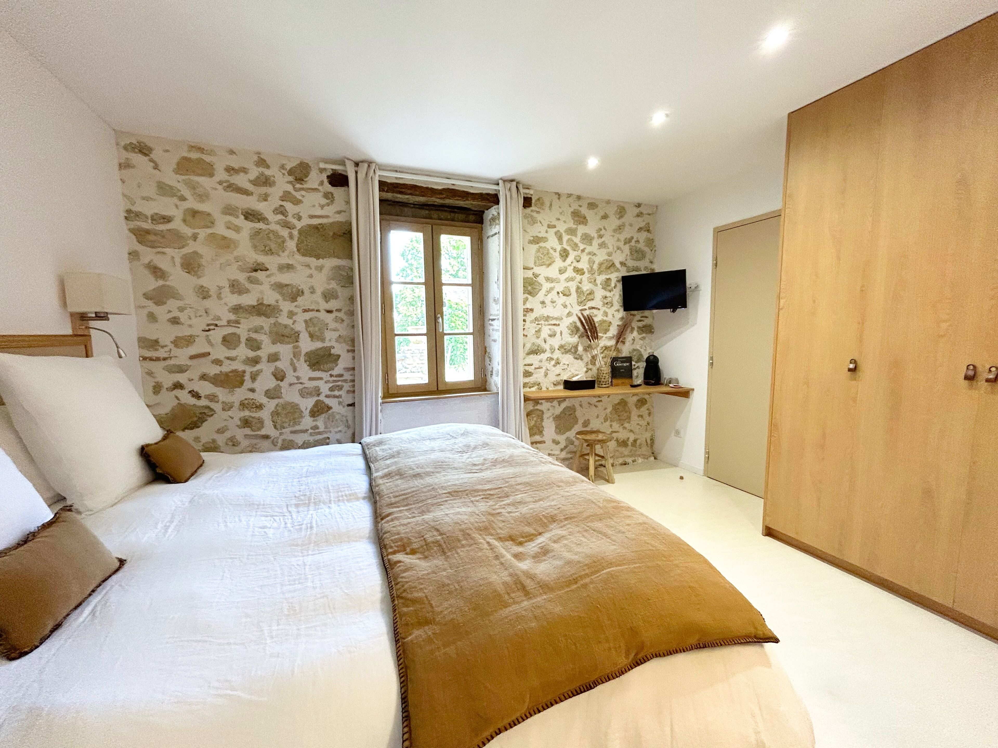 A bedroom with a large bed, a wooden wardrobe, a television on the wall, and a window with curtains.