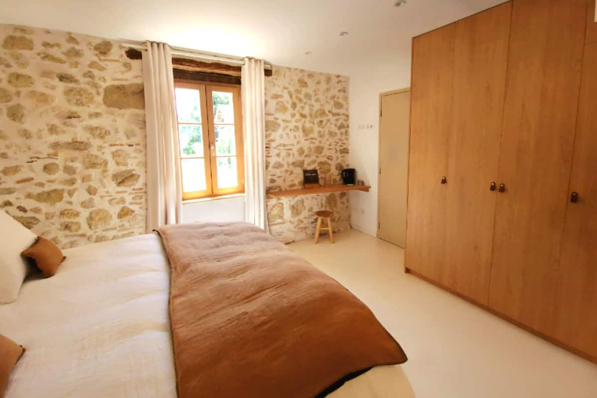 A bedroom with a stone wall, a large wooden wardrobe, a bed with a brown blanket, and a window with curtains.