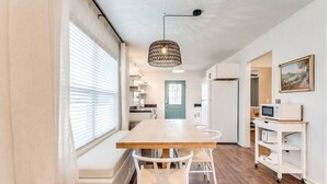A modern kitchen with a wooden table and white chairs, a hanging light fixture, and a view of the adjacent living area.