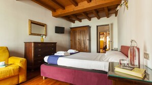 Quarto com teto de madeira, cama com colcha branca e roxa, cómoda de madeira, poltrona amarela e mesa de vidro com um livro e um copo.
