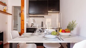 Uma área de refeições com uma mesa branca posta para quatro pessoas, rodeada por cadeiras brancas. Há uma cesta de frutas na mesa, incluindo uvas, uma abacaxi e uma maçã vermelha. O espaço possui armários de madeira escura acima do balcão e um revestimento de azulejos brancos.