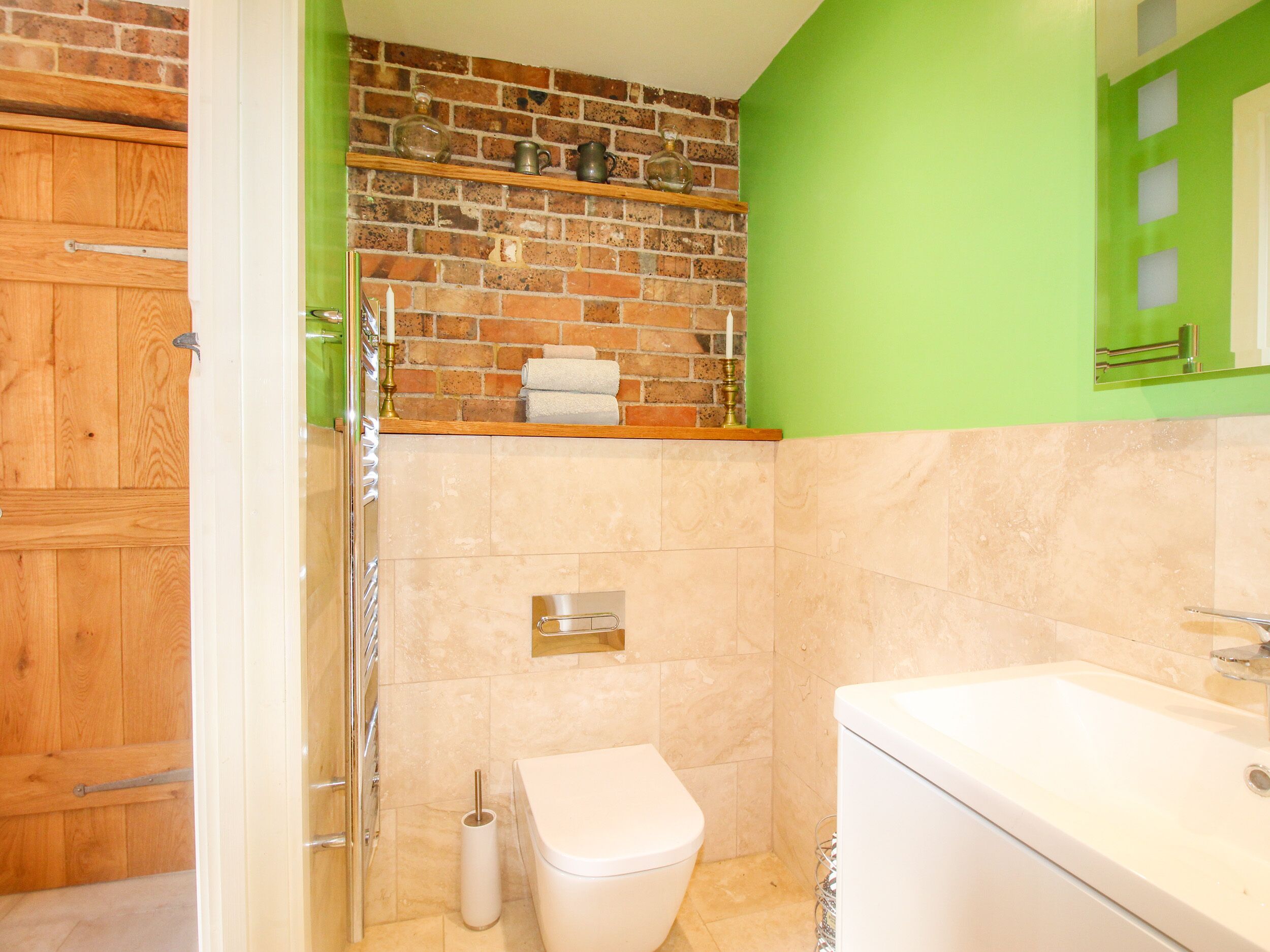 A bathroom with a brick wall, a green wall, a wooden door, a white toilet, a white sink, and a towel rack with towels.