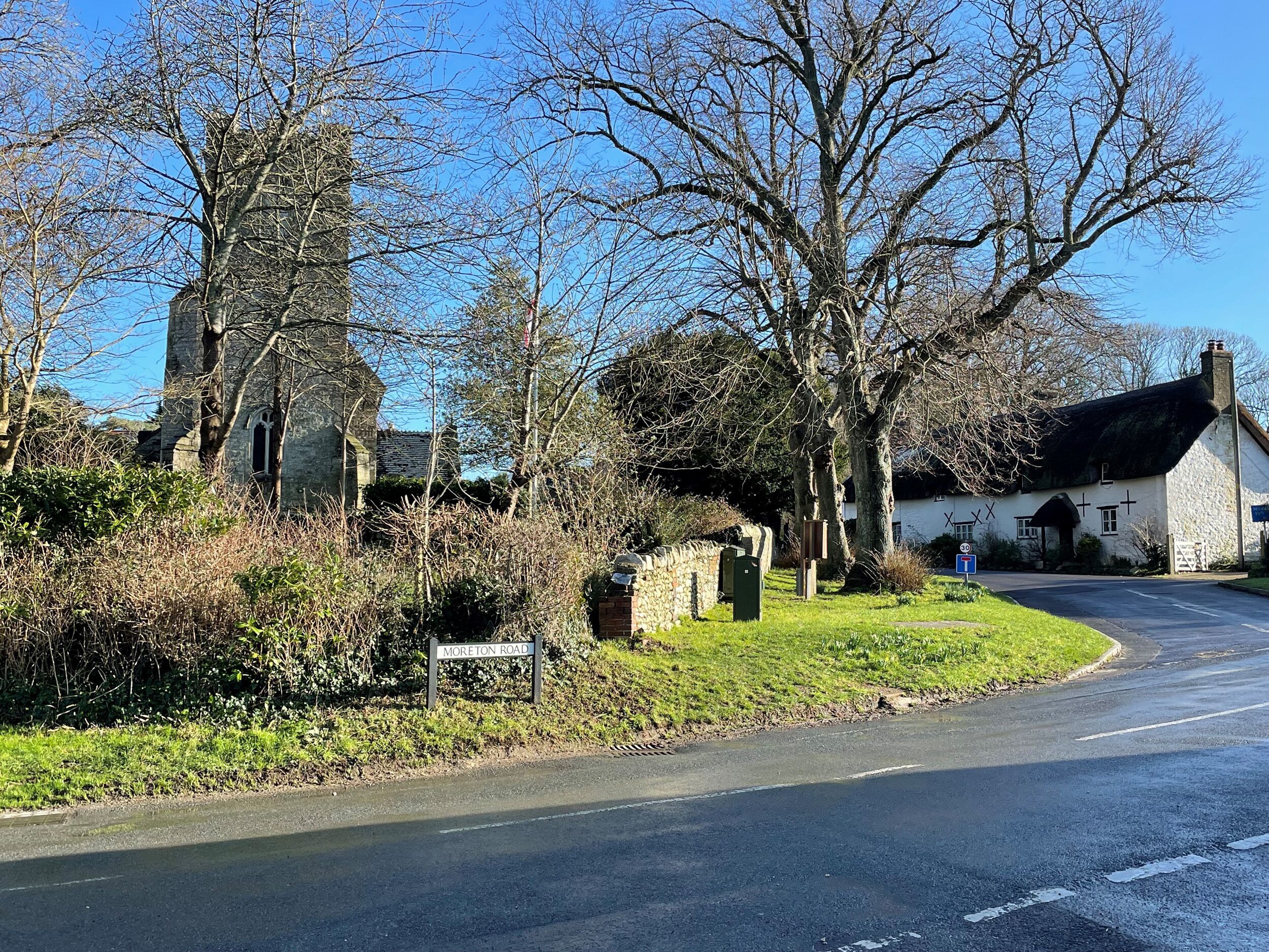 Uma estrada rural com uma igreja de pedra, uma casa branca e uma placa indicando Moreton Road.
