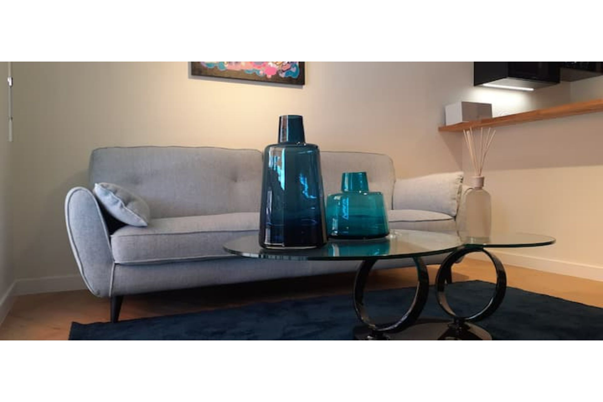 Una sala de estar con un sofá gris, una mesa de centro de vidrio con jarrones azules y un cuadro en la pared.