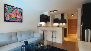 A modern living room with a sofa, a coffee table, and a painting on the wall. There is a kitchen area with a counter and stools. - Allées du Parc, high-end apartment 🌳🌲🌾 (Dijon)
