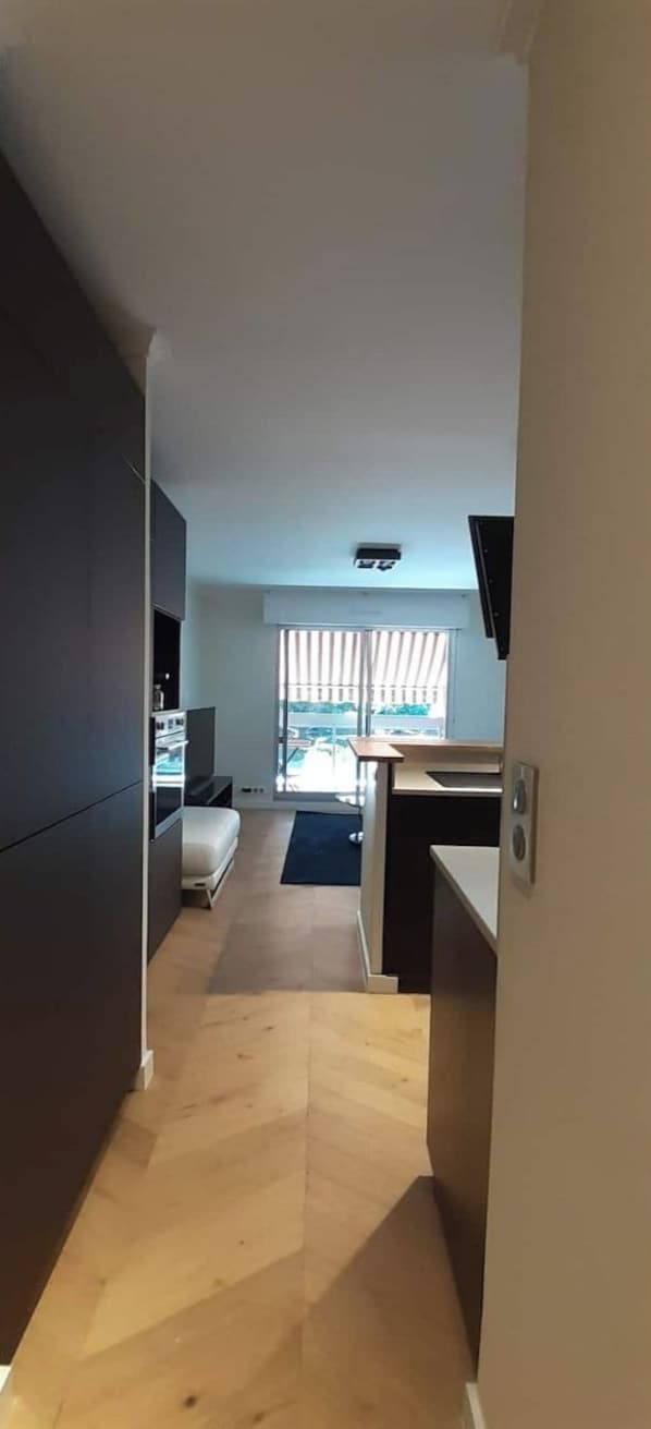 A narrow hallway leading to a living area with a sofa, a TV, and a window with blinds.