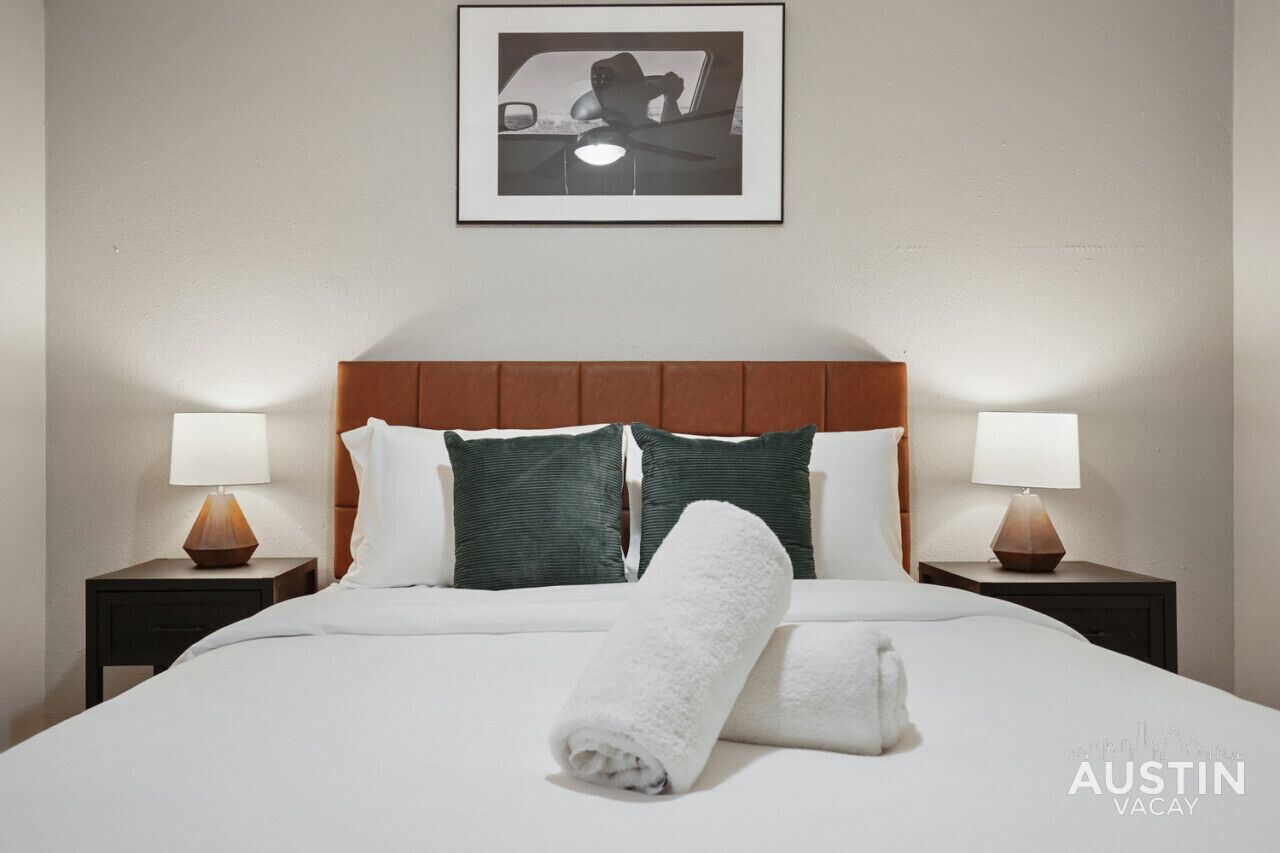 A neatly made bed with a white comforter, two green pillows, and a rolled white towel. There are two bedside tables with lamps, and a framed picture hangs above the bed.