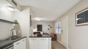 A modern kitchen with white cabinets, a black countertop, and a built-in oven. There is a window with blinds, a plant, and a framed artwork on the wall.