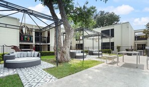 An outdoor seating area with round tables and sofas, a large tree, and a building with a white facade and black-framed windows. - Chelsea #249 xxxxxxx (Austin)