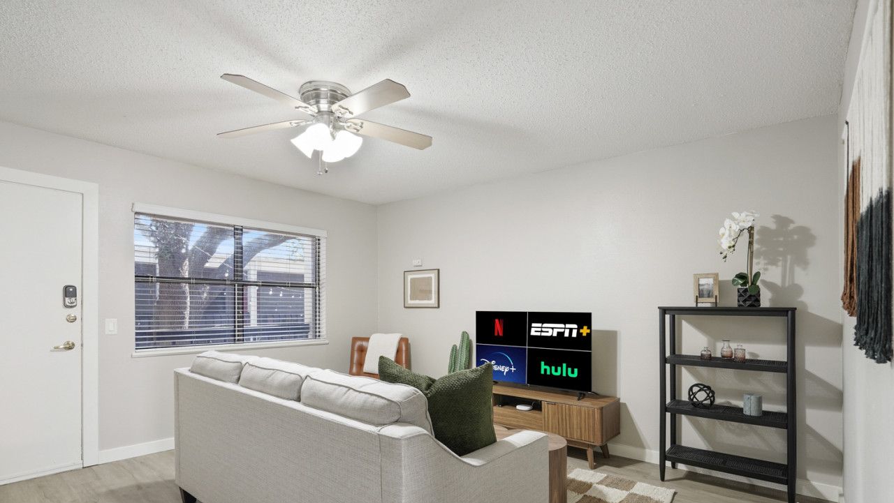 Een woonkamer met een grijze bank, een plafondventilator, een tv waarop streamingdiensten worden getoond, een houten bijzettafeltje en een zwart kastenrek.