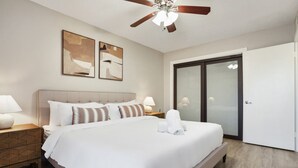 A hotel room with a bed, a ceiling fan, two wall art pieces, a nightstand, a lamp, a bathroom with a glass door, and a wooden floor. - Chelsea #211 xxxxxxx (Austin)