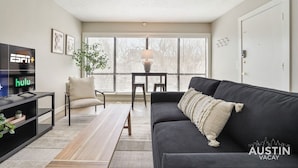 A living room with a black sofa, a wooden coffee table, a beige armchair, a TV displaying ESPN and Hulu, and a large window with blinds. - Chelsea #211 xxxxxxx (Austin)