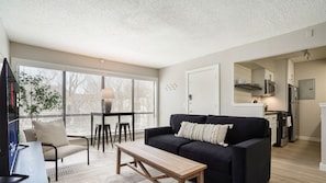 A modern living room with a black sofa, a wooden coffee table, and a TV on a stand. There is a dining area with a table and chairs, and a kitchen in the background.