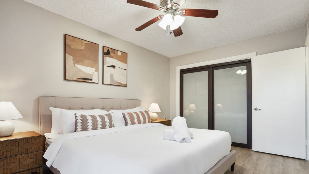 Habitación de hotel con cama, ventilador de techo, dos cuadros en la pared, mesita de noche, lámpara, baño con puerta de vidrio y suelo de madera.