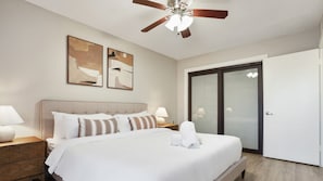 A hotel room with a bed, a ceiling fan, two wall art pieces, a nightstand, a lamp, a bathroom with a glass door, and a wooden floor.
