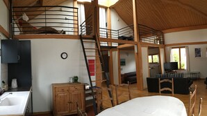 Un vaste espace de vie de style loft avec un plafond haut, des poutres apparentes, un lit superposé, un coin cuisine et un espace détente avec un téléviseur.