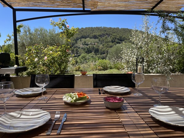 A table set for two with plates, cutlery, and glasses, overlooking a scenic landscape with trees and hills.