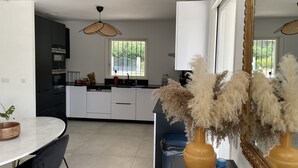 A modern kitchen with white cabinets, a black countertop, and a window with a view of greenery. There is a dining area with a round table and chairs, and a vase with dried plants on the counter. - Pretty villa in the Bandol vineyards, 15 minutes from the beaches. (Le Beausset)