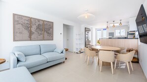 Ein modernes Wohnzimmer mit einem hellblauen Sofa, einem Essbereich mit rundem Tisch und Stühlen sowie einer Küche mit Kühlschrank und einer Pendelleuchte.