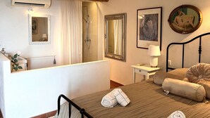 Ein Schlafzimmer mit Holzboden, einem Bett mit Kissen, einem Nachttisch mit Lampe, einem großen Spiegel und einem Badezimmer mit Dusche und Toilette.