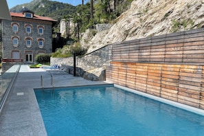A swimming pool with a stone wall and wooden paneling, surrounded by a stone building and a mountainous backdrop. - Modern House Apartments (Menaggio)