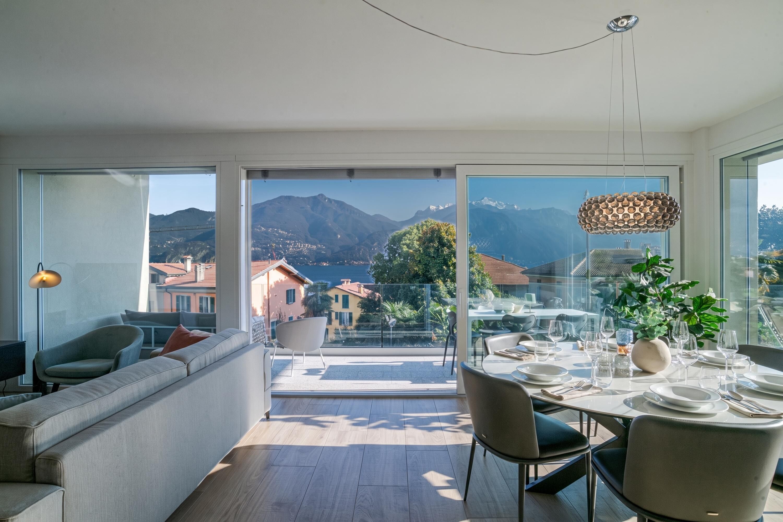 A modern living room with a sofa, a dining area with a table set for four, and a balcony with a mountain view.
