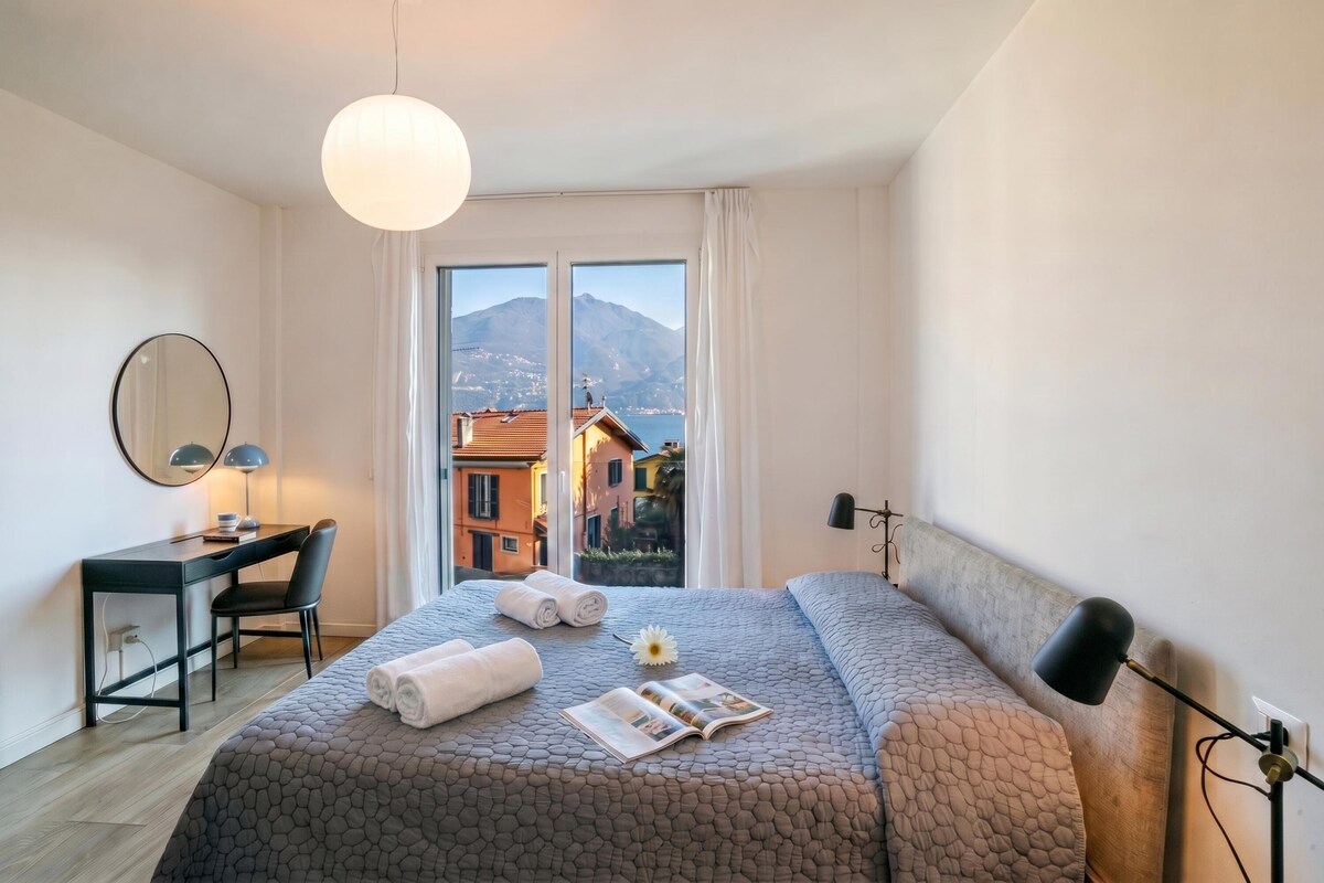 A hotel room with a bed, a desk, a chair, a lamp, and a view of buildings and mountains through the window.