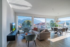 A modern living room with a sofa, chairs, a TV, and a dining area with a view of mountains. - Modern House Apartments (Menaggio)