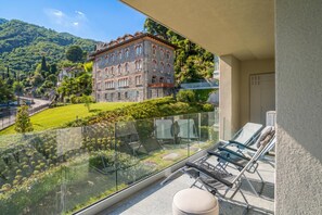 A balcony with a glass railing overlooking a mountainous landscape, a stone building, and a clear sky.