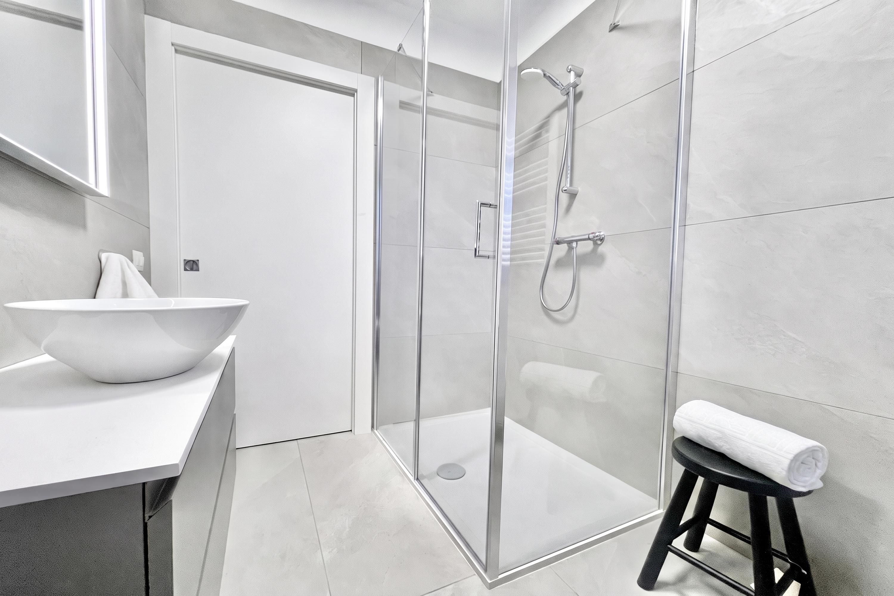 Une salle de bain moderne avec une douche à parois de verre, un lavabo blanc en forme de cuvette et un tabouret noir sur lequel est posée une serviette.
