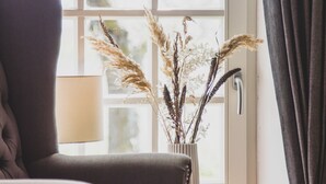 Ein Zimmer mit einem dunkelbraunen Polsterarmstuhl, einem Fenster mit Blick auf Bäume und einer Vase mit getrockneten Pflanzen.