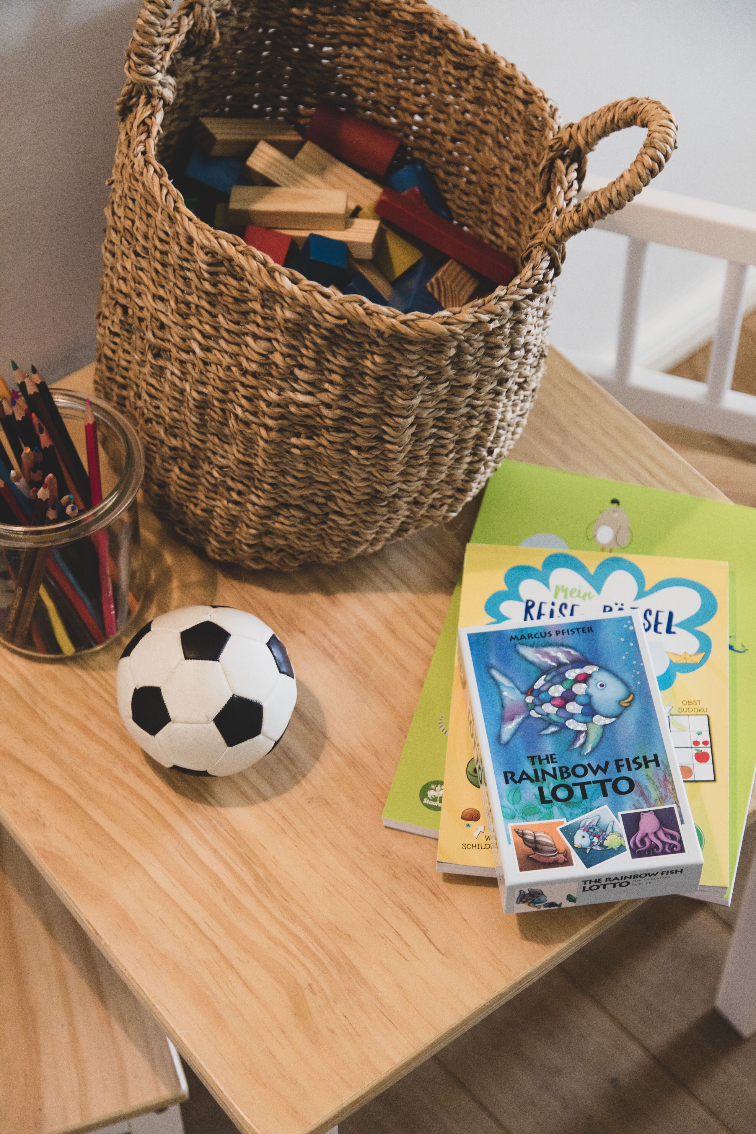 A wooden table with a woven basket, a soccer ball, a jar of pencils, and a book titled "The Rainbow Fish Lotto".