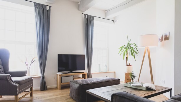 Ein modernes Wohnzimmer mit Sofa, Hocker und Couchtisch. Ein Fernseher steht auf einem Lowboard, eine Stehlampe sorgt für Licht und eine Topfpflanze bringt Leben in den Raum.