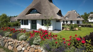 A thatched-roof cottage with a white exterior, surrounded by a well-maintained garden with various flowering plants and a stone wall. - Ferienhaus Seeadler 34 - Der Natur Ganz nah (Fuhlendorf)