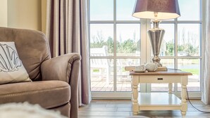 A cozy living room with a brown leather sofa, a white side table with a lamp, and a view of a deck and outdoor area through large windows. - Ferienhaus Haubenlerche 20 - Der Natur Ganz nah (Fuhlendorf)